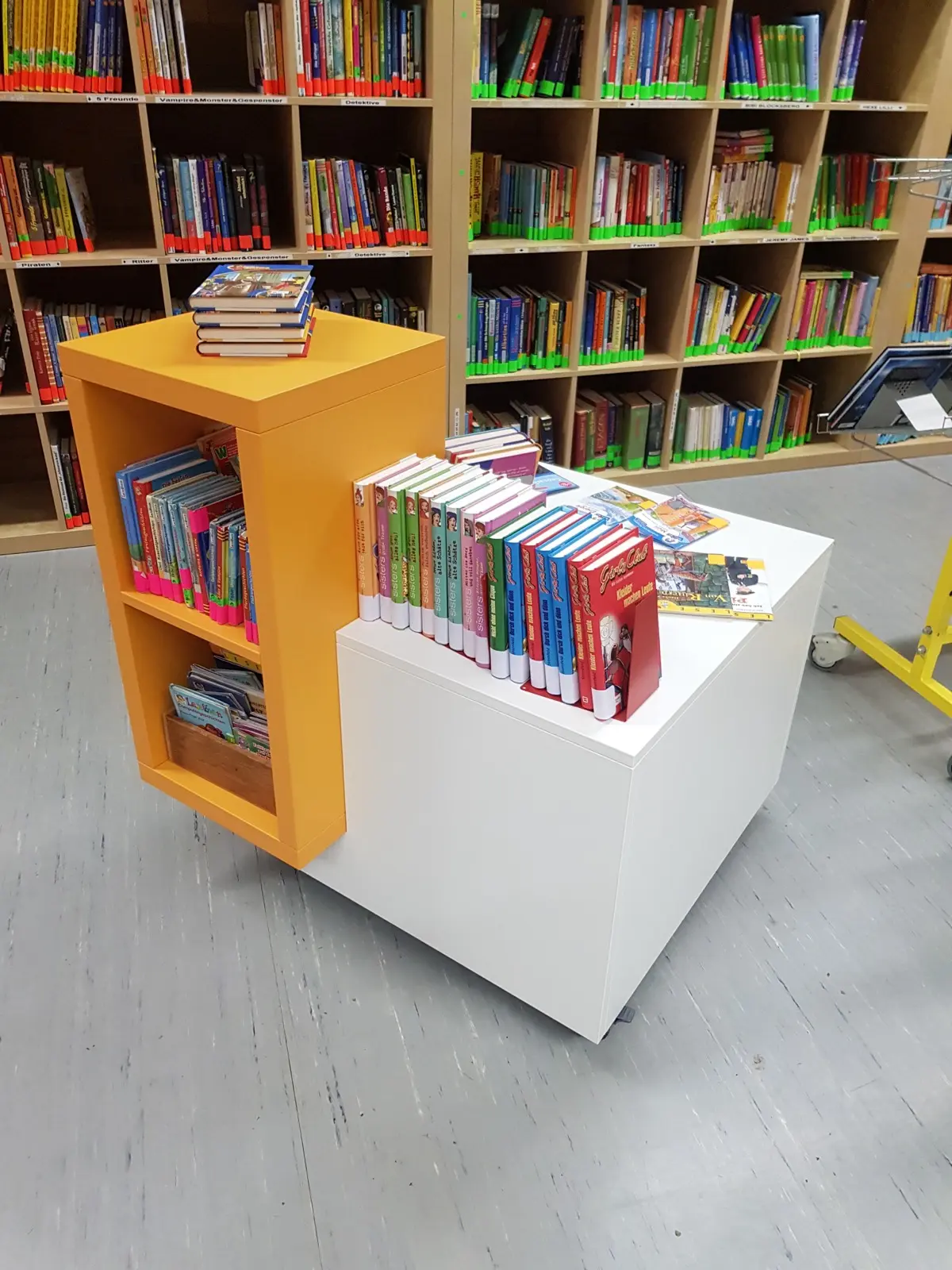 Ein Bücherwagen mit bunten Kinderbüchern steht vor gefüllten Regalen der Bücherei