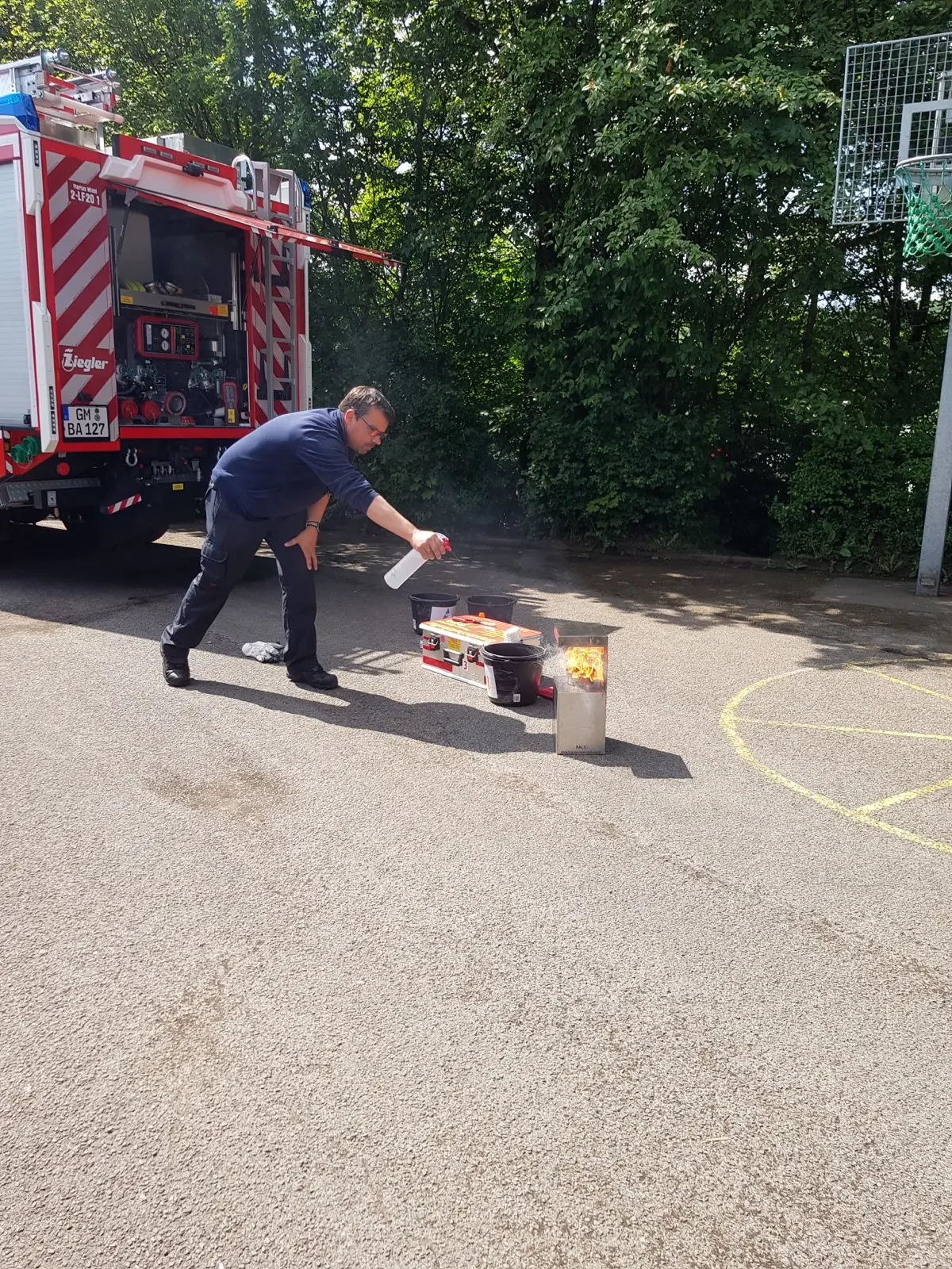 Ein Feuerwehrmann löscht auf dem Schulhof ein kleines Übungsfeuer vor dem Löschfahrzeug