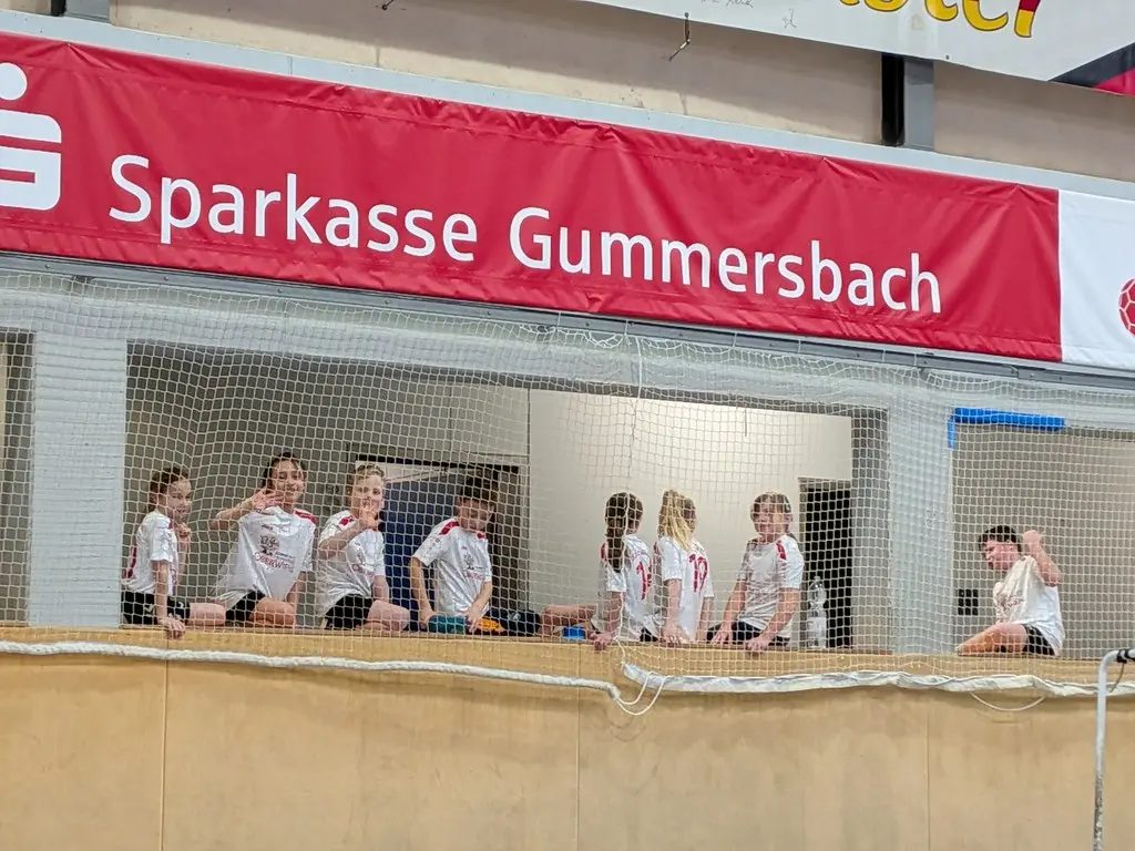 Kinder in Schultrikots sitzen unter einem Sparkassen-Banner auf der Tribüne