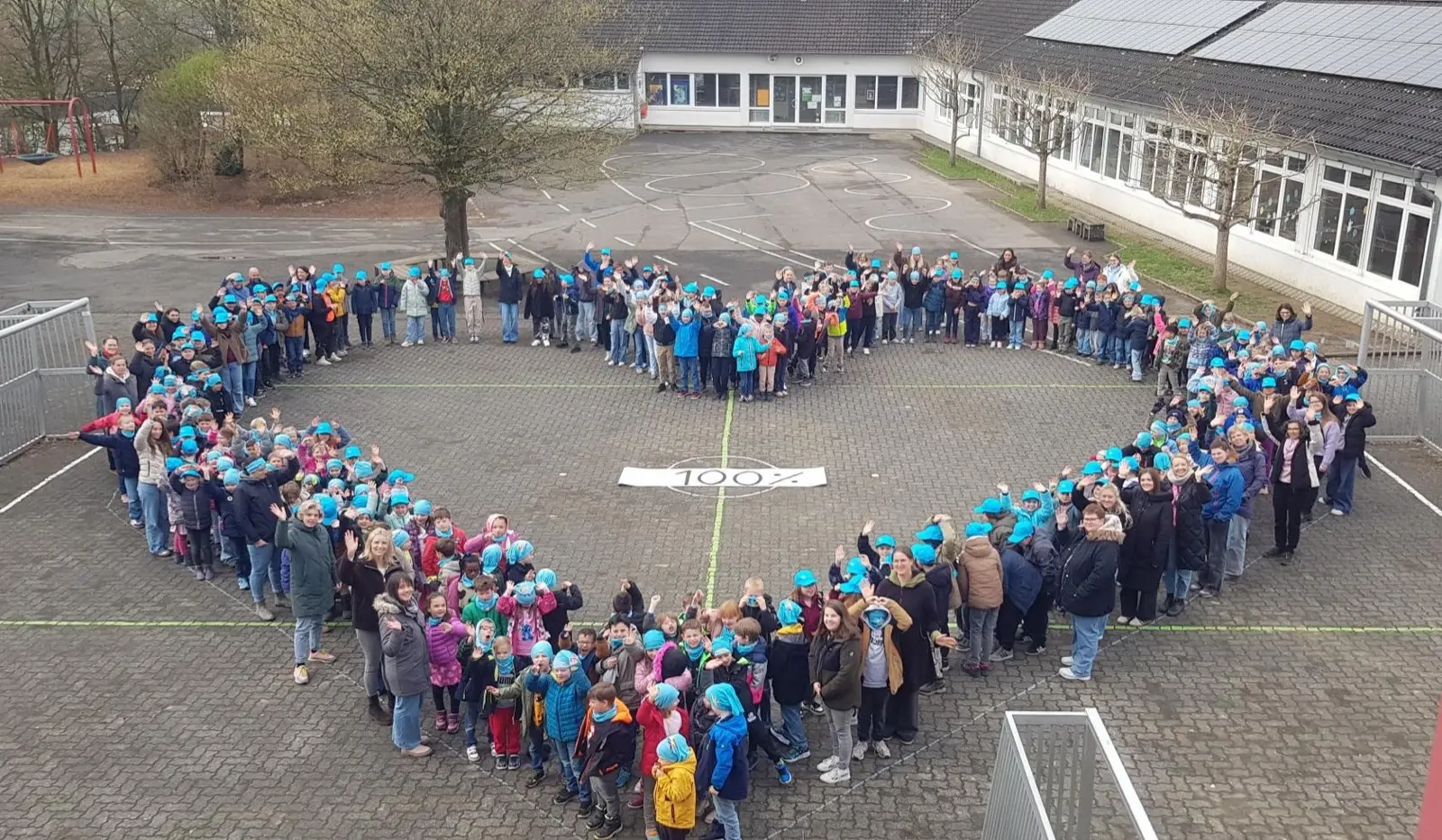 Die ganze Schulgemeinschaft bildet auf dem Schulhof mit blauen Mützen ein großes Herz