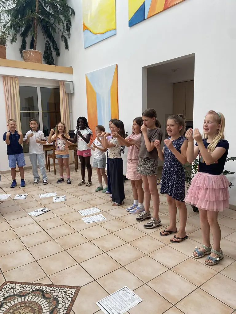 Eine Gruppe Kinder singt in einer Reihe im hellen Atrium und begleitet das Lied mit Handgesten