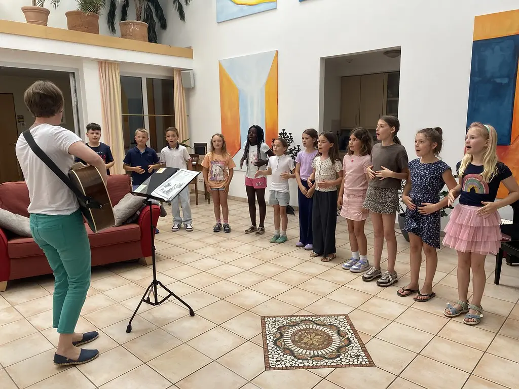 Eine Lehrerin mit Gitarre steht am Notenpult vor dem singenden Kinderchor im Atrium