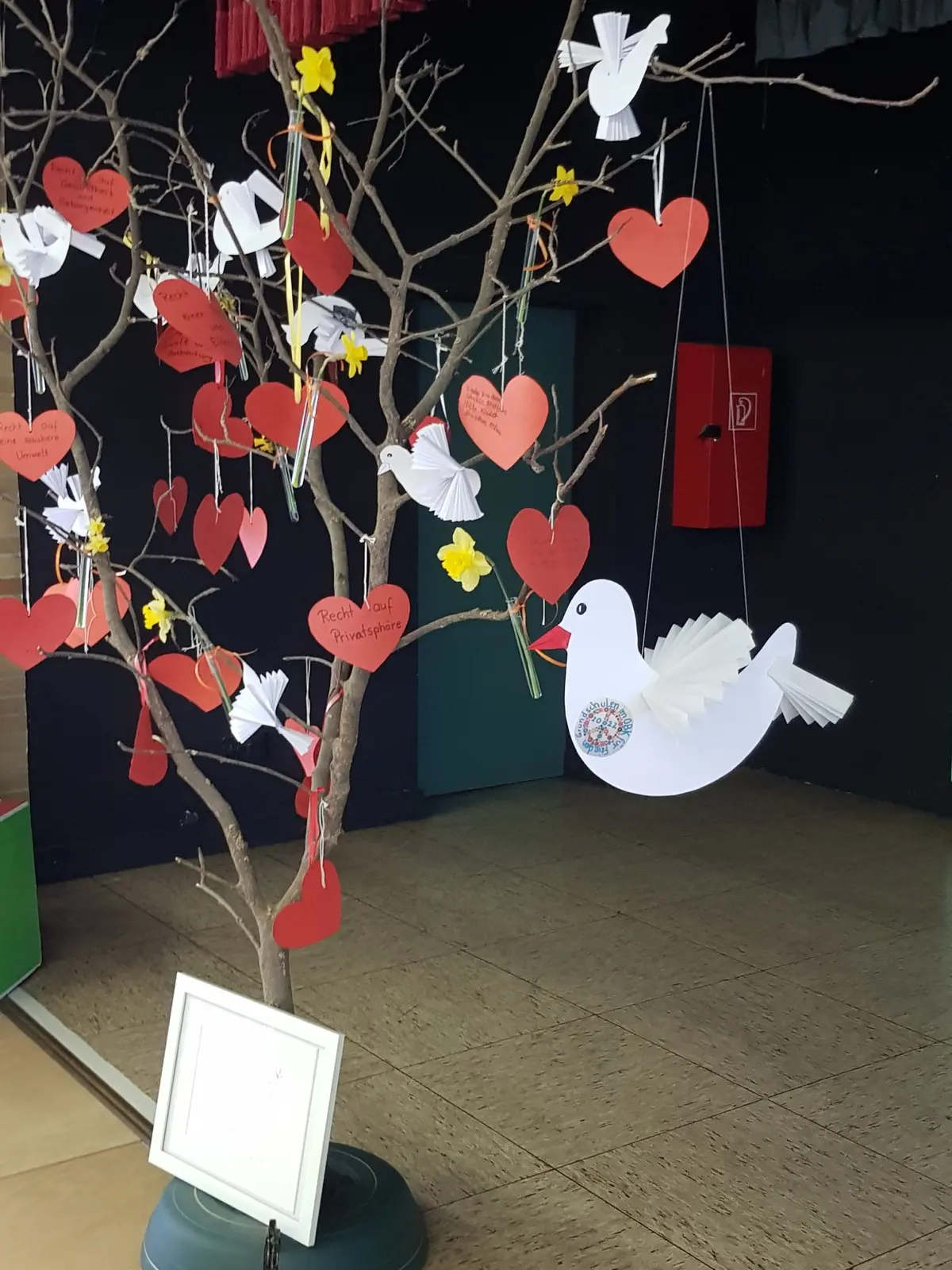 Ein Ast mit roten Herzen, beschrifteten Karten und Papiertauben als Kinderrechte-Baum in der Aula