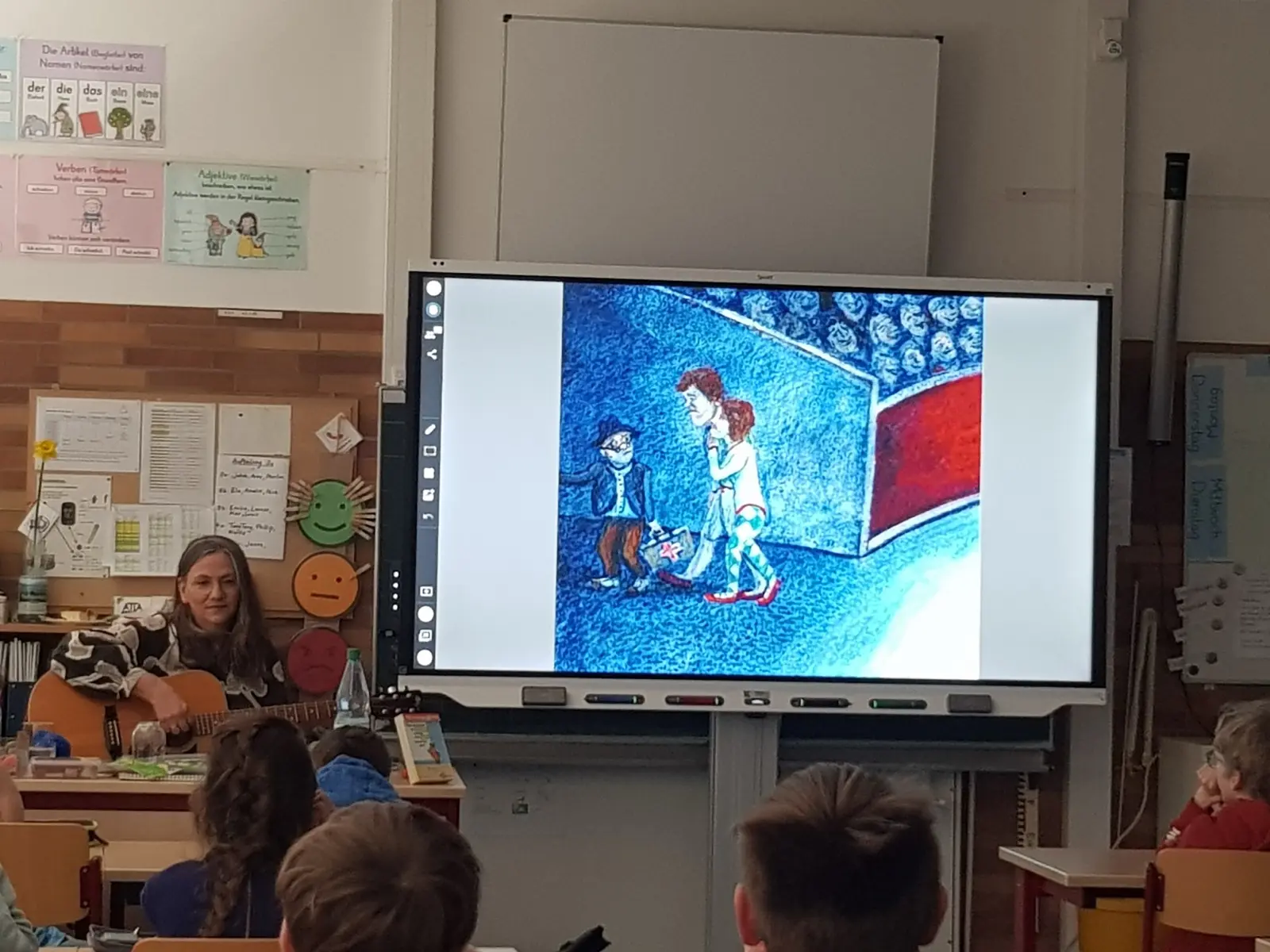 Eine Lehrerin begleitet mit der Gitarre eine Bilderbuchgeschichte auf dem Whiteboard im Klassenraum