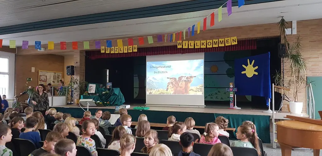 Kinder sitzen in der festlich geschmückten Aula vor der Bühne mit Leinwand