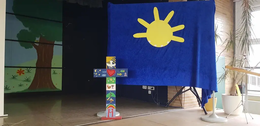 Buntes, von Kindern bemaltes Holzkreuz vor blauem Tuch mit gelber Sonne