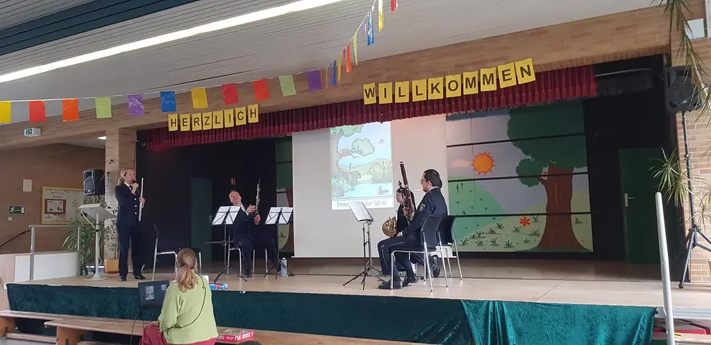 Musizierende Erwachsene mit Flöte und Fagott auf der Bühne unter dem Schriftzug „HERZLICH WILLKOMMEN“