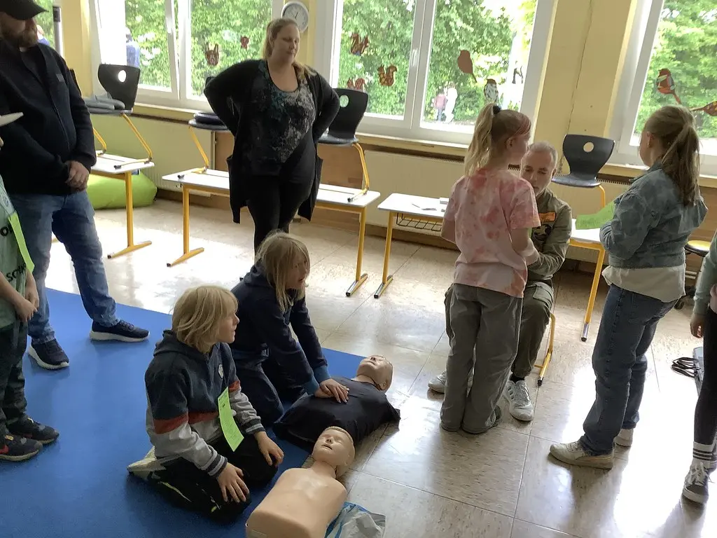 Kinder üben Herzdruckmassage an Übungspuppen, Erwachsene leiten sie an
