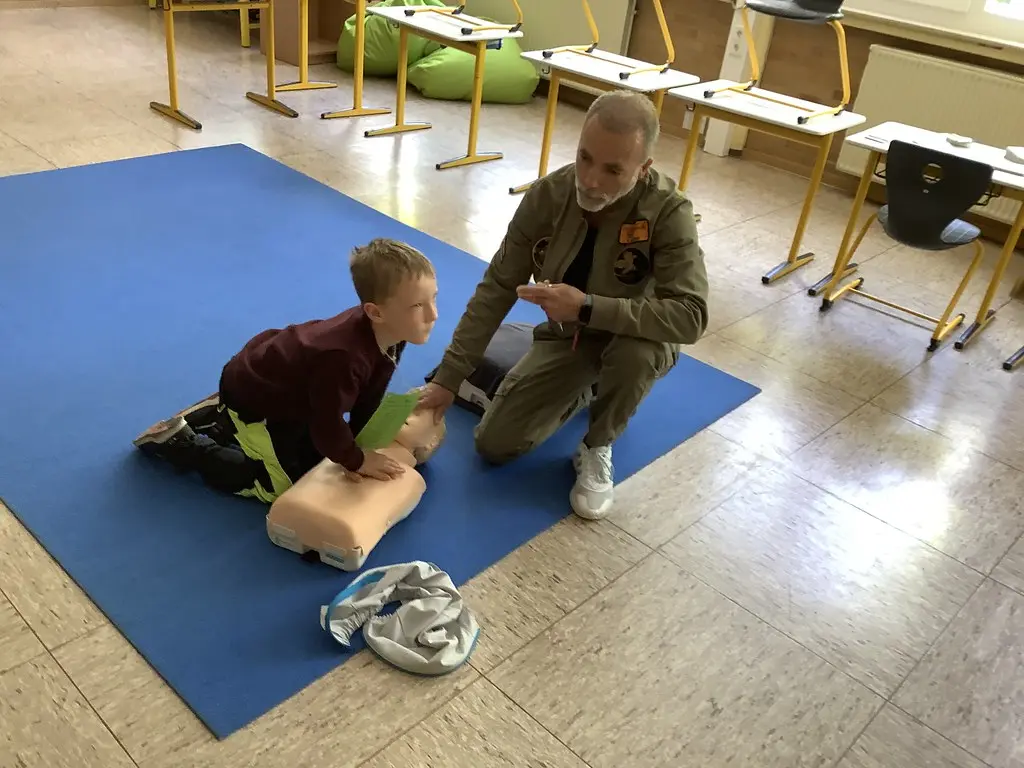 Ein Kind übt Herzdruckmassage an einer Übungspuppe, ein Erwachsener hilft