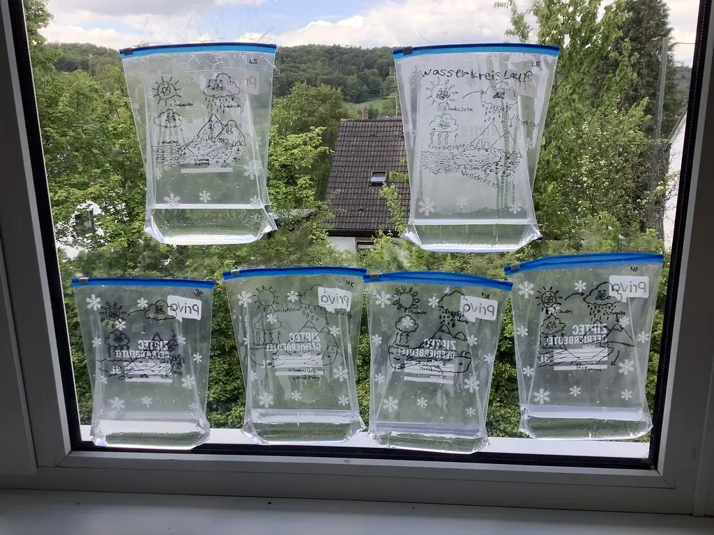 Beschriftete Gefrierbeutel mit Wasserkreislauf-Zeichnungen kleben an einer Fensterscheibe