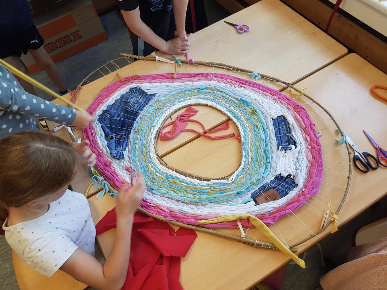 Mehrere Kinder weben gemeinsam ein großes rundes Bild aus Stoffstreifen